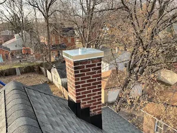 A brick chimney with a metal cap on an asphalt-shingle roof, bare winter trees behind