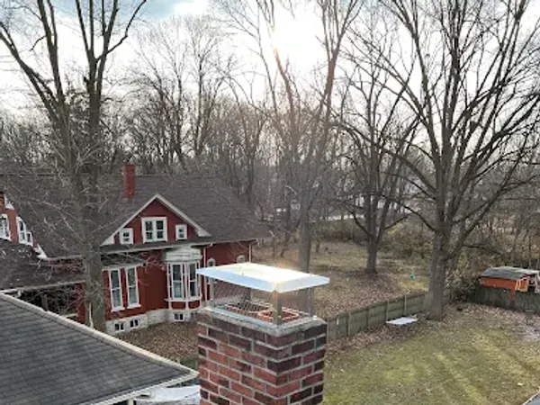 A brick chimney and crown on a shingle roof, a neighborhood of older homes behind