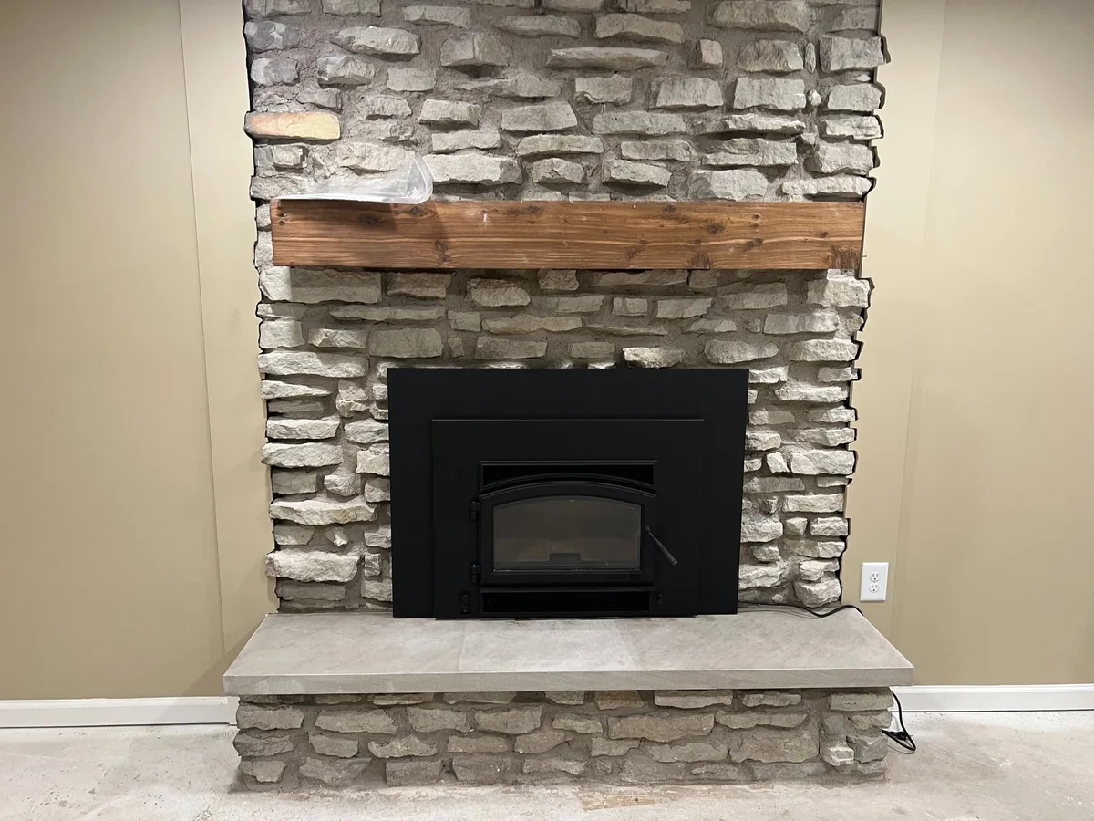 A wood-burning insert in a rough stone fireplace with a timber mantel