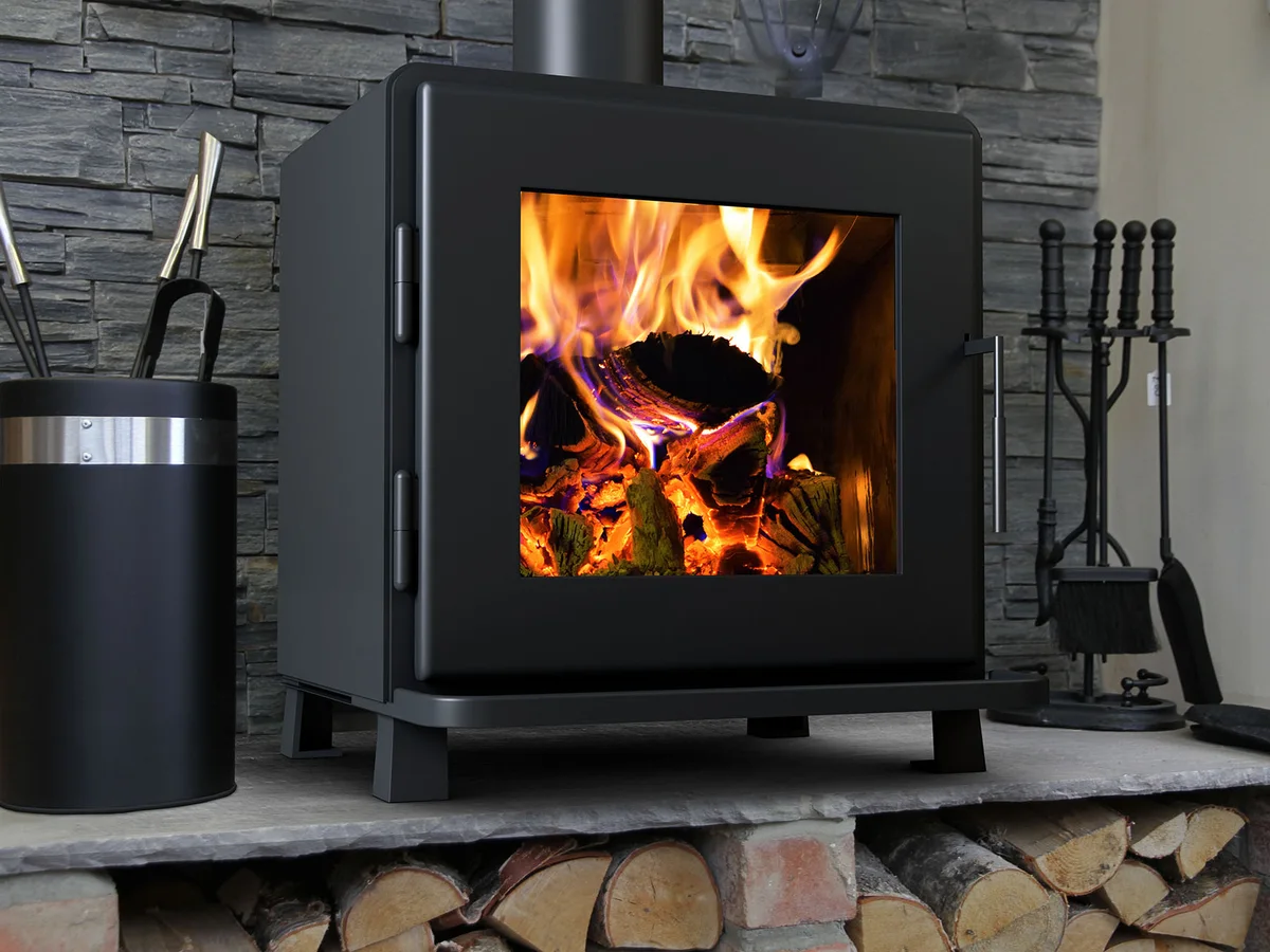 A wood stove burning on a stone hearth, split logs stacked beneath it